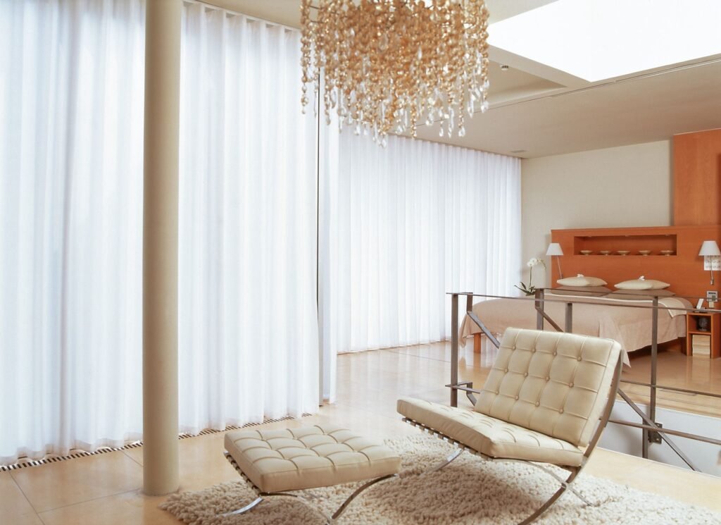 Sheer curtains falling from a ceiling track beside a lounge chair and chandelier